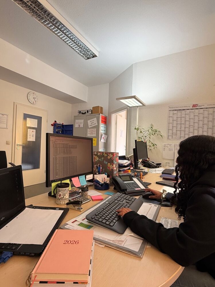 Eine Person sitzt an einem Büroarbeitsplatz und arbeitet konzentriert an einem Computer. Der Schreibtisch ist mit verschiedenen Büromaterialien wie Tastatur, Maus, Notizblöcken, Stiften, einem Telefon, Ordnern, einem Kalender für das Jahr 2026 und persönlichen Gegenständen ausgestattet. Im Hintergrund sind ein Wandkalender und ein Regal mit weiteren Utensilien zu sehen.