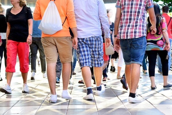 Mehrere Personen gehen auf einem sonnigen, gepflasterten Platz. Die Aufnahme zeigt die Szene aus niedriger Perspektive von hinten. Die Menschen tragen sommerliche Kleidung wie kurze Hosen, T-Shirts und leichte Schuhe. Sonnenlicht wirft geometrische Schattenmuster auf den Boden.
