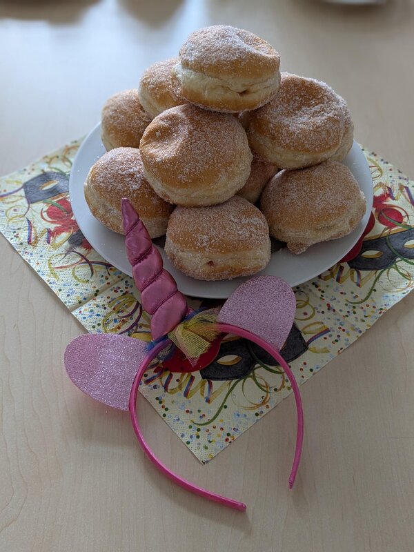 Ein Teller mit einer großen Portion Berliner (Krapfen) mit Puderzucker liegt auf einem farbenfrohen Karnevals-Servietten-Set. Daneben liegt ein pinker Haarreif mit einem Einhorn-Horn und glitzernden Ohren, passend zur Faschingsstimmung.