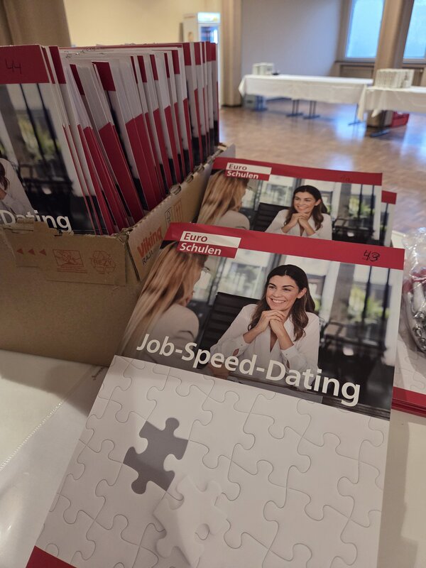 Mehrere Broschüren der Euro-Schulen zum Job-Speed-Dating liegen in einem Karton und auf einem Tisch zur Ausgabe an die Teilnehmenden.