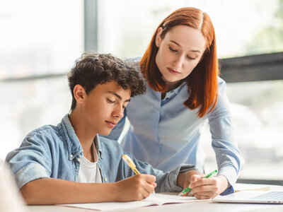 Eine Frau mit langen roten Haaren und einem hellblauen Hemd steht hinter einem jungen Mann mit kurzem, lockigem Haar und einem blauen Denim-Hemd. Beide sind an einem Tisch und scheinen sich auf ein Notizbuch zu konzentrieren, in das der junge Mann schreibt. Die Frau beobachtet und unterstützt ihn dabei. Auf dem Tisch liegt auch ein Laptop.