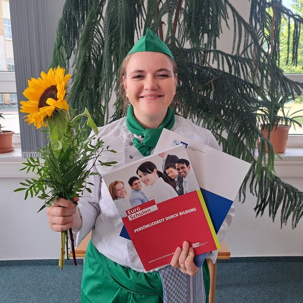 Eine Absolventin grinst zufrieden in die Kamera. Sie trägt ein Kochoutfit mit grüner Schürze, Halstuch und Kochmütze. In der linken Hand hält sie die Prüfungszeugnisse in Mappen der IHK und der Euro-Schulen, in der rechten Hand einen Strauß mit einer Sonnenblume.