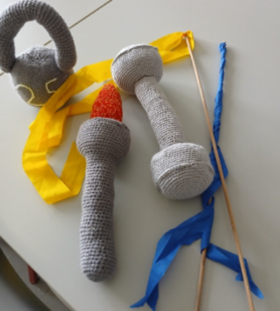 Gehäkelte Sportgeräte auf einem Tisch: eine Kettlebell, zwei Hanteln sowie gelbe und blaue Gymnastikbänder an Stäben.