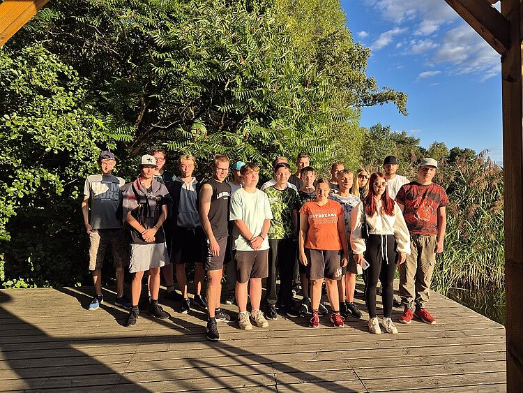 Gruppenfoto von Jugendlichen auf einer Holzplattform vor dichtem Grün, aufgenommen bei Sonnenschein.
