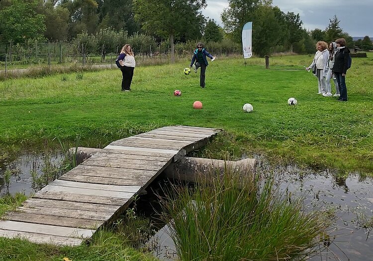 Team-Building beim Fußballgolf Fünf Frauen mit fünf Fußbällen stehen auf einer grünen Wiese. Eine Frau versucht, den Fußball in Richtung eines Tümpels über eine kleine Holzbrücke zu schießen. Im Hintergrund steht eine Beachflag, die vermutlich Werbung für den Fußballgolf-Standort macht.