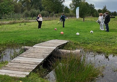 Team-Building beim Fußballgolf Fünf Frauen mit fünf Fußbällen stehen auf einer grünen Wiese. Eine Frau versucht, den Fußball in Richtung eines Tümpels über eine kleine Holzbrücke zu schießen. Im Hintergrund steht eine Beachflag, die vermutlich Werbung für den Fußballgolf-Standort macht.
