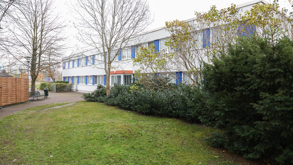 Rasenfläche, Büsche und Bäume vor einem zweigeschossigen, modernen Gebäudekomplex mit weißer Fassade und blauen Fensterrahmen