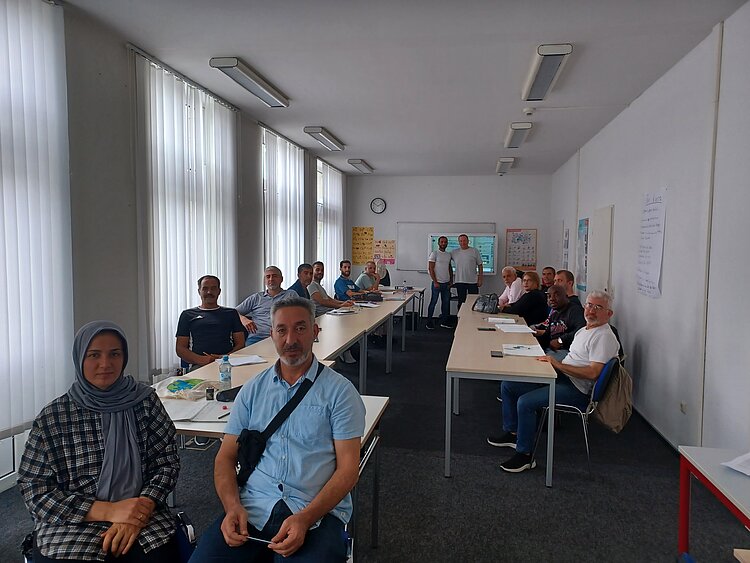 Eine Gruppe von etwa fünfzehn Erwachsenen sitzt in einem Klassenraum an langen Tischen und blickt zur Kamera. Im Hintergrund stehen zwei Personen vor einer Tafel. Auf den Tischen liegen Hefte, Stifte und Wasserflaschen, an den Wänden hängen Poster und Flipchart-Papiere.
