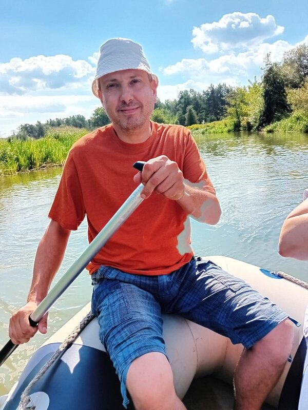 Ein Mann mit T-Shirt, kurzer Jeans und einem hellen Sonnenhut aus Stoff sitzt auf dem Rand eines Schlauchboots mit einem Ruder in der Hand. Dahinter sieht man den kleinen Fluss mit begrüntem Ufer und einige Schönwetterwolken am ansonsten blauen Himmel.