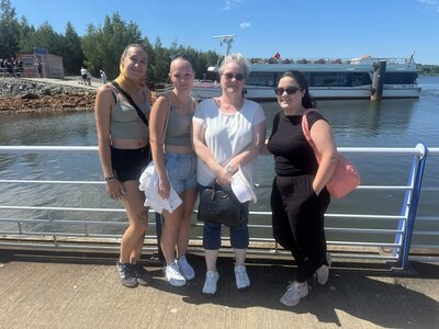 Vier Frauen posieren bei sonnigem Wetter an einem Geländer vor einem Gewässer; im Hintergrund liegt ein Ausflugsschiff am Ufer.