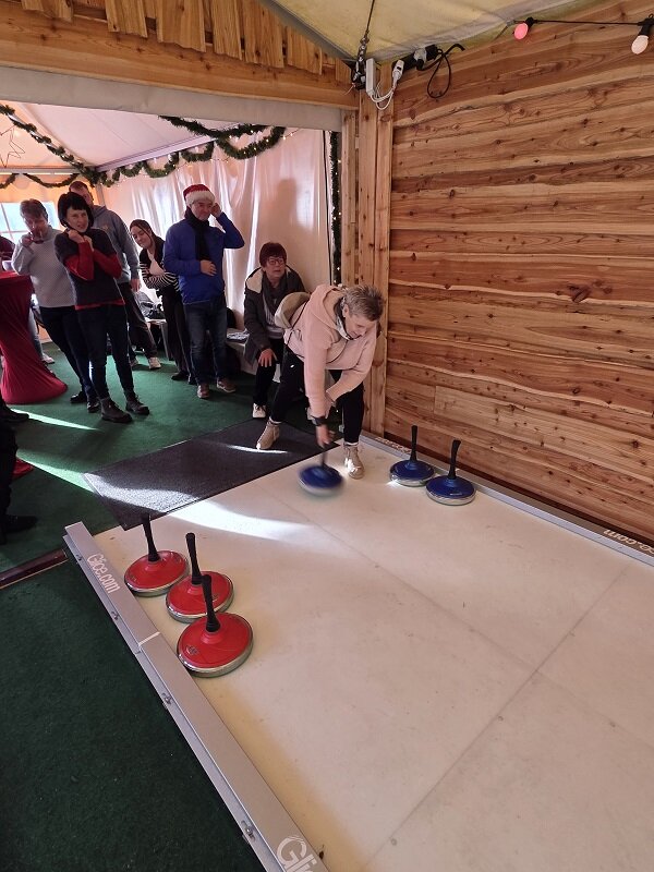 Eine Frau stößt einen blauen Eisstock über die Bahn, während mehrere Personen im Hintergrund zuschauen und warten.