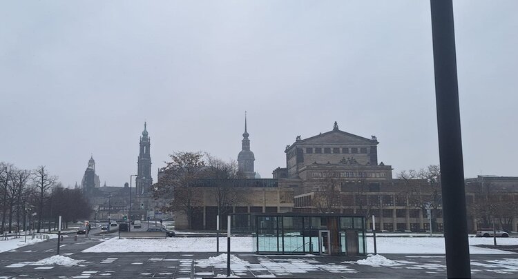 Blick auf einen verschneiten, weitläufigen Platz in Dresden mit Straßen und wenigen Autos im Vordergrund, dahinter mehrere historische Gebäude mit Türmen unter einem grauen Winterhimmel.