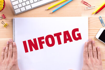 Die Aufschrift "Infotag" in großen roten Lettern steht auf einem Zeichenblock geschrieben, der auf einem Holztisch liegt und neben dem rechts und links die Hände einer Person flach aufliegen. Um den Block verteilt liegen Stifte, Büroklammern und ein Handy und es ist die Ecke einer Tastatur zu sehen.