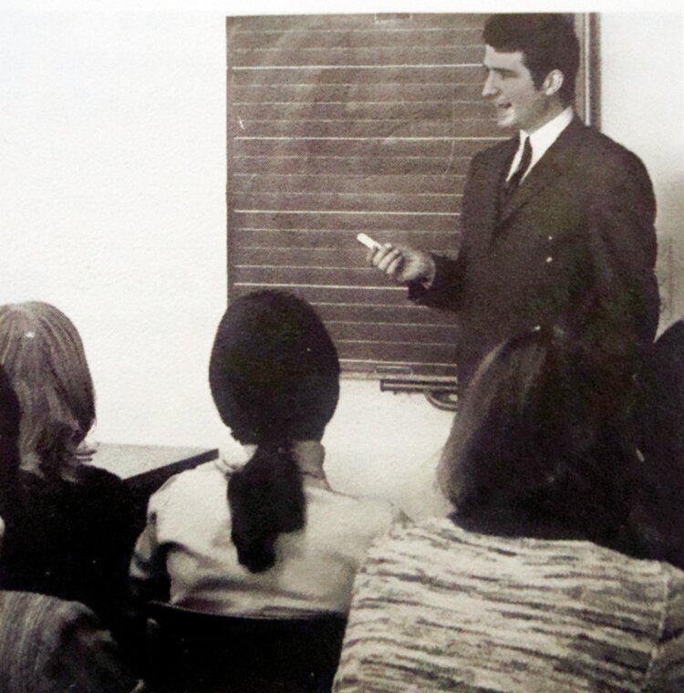 Ein Mann in Anzug steht vor einer Tafel und hält einen Kreidestift in der Hand, während er zu einer Gruppe von Personen spricht, die mit dem Rücken zur Kamera sitzen. Das Bild hat einen sepiafarbenen Ton und wirkt wie eine historische oder alte Aufnahme.