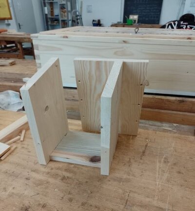 Eine Holzwerkbank in einer Werkstatt mit einem unfertigen Nistkasten aus hellem Holz. Das Nistkasten-Gerüst besteht aus einer Bodenplatte und drei senkrechten Platten, die als Seitenwände und Rückwand dienen. Im Hintergrund sind weitere Holzbretter und Werkzeuge sichtbar.