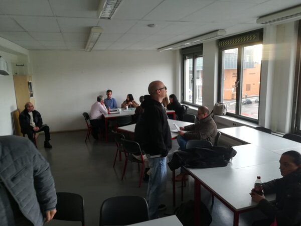 Teilnehmende in einem Schulungsraum, einige im Gespräch am Tisch, eine Person steht im Vordergrund; Tageslicht durch die Fensterfront.