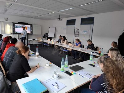 Große Seminargruppe sitzt an U-förmig gestellten Tischen; vorne steht ein Referent neben einem Bildschirm, Teilnehmende hören zu und machen Notizen.