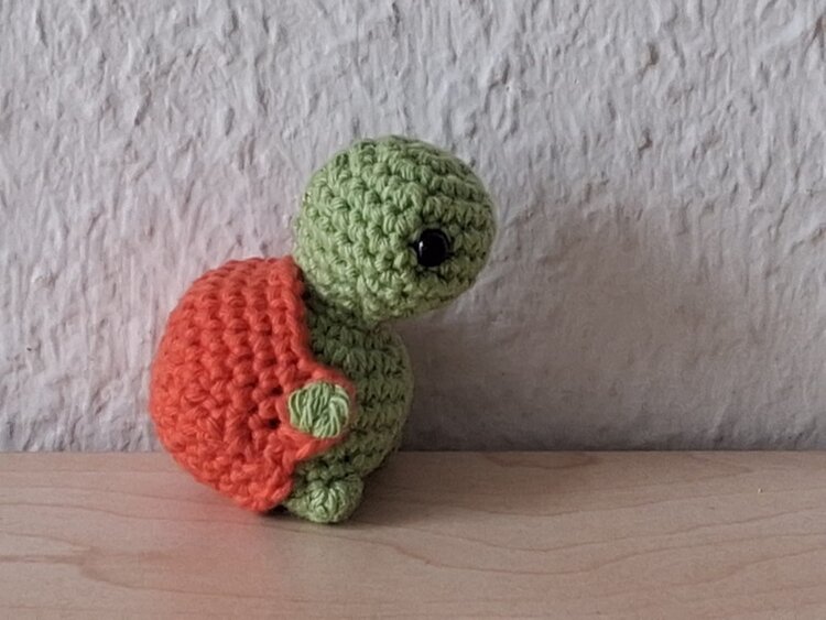 Gehäkelte Schildkröte mit grünem Kopf und Beinen sowie einem orangefarbenen Panzer steht auf einer hellen Holzoberfläche vor einer strukturierten, hellgrauen Wand. Das Auge ist als schwarzer Knopf gestaltet.