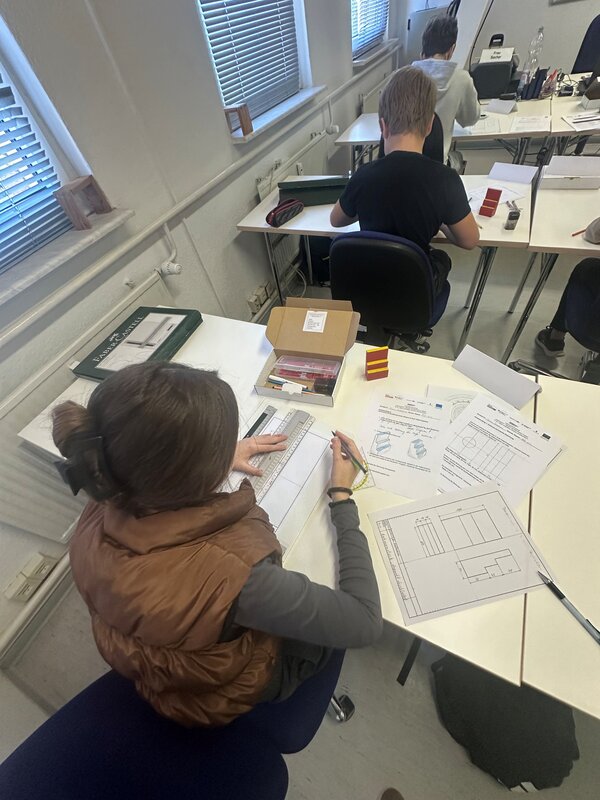 Eine Schülerin erstellt mit Lineal und Stift eine technische Zeichnung. Auf dem Tisch liegen Arbeitsblätter mit Maßzeichnungen, Zeichenmaterialien und kleine selbst gefertigte Modelle.
