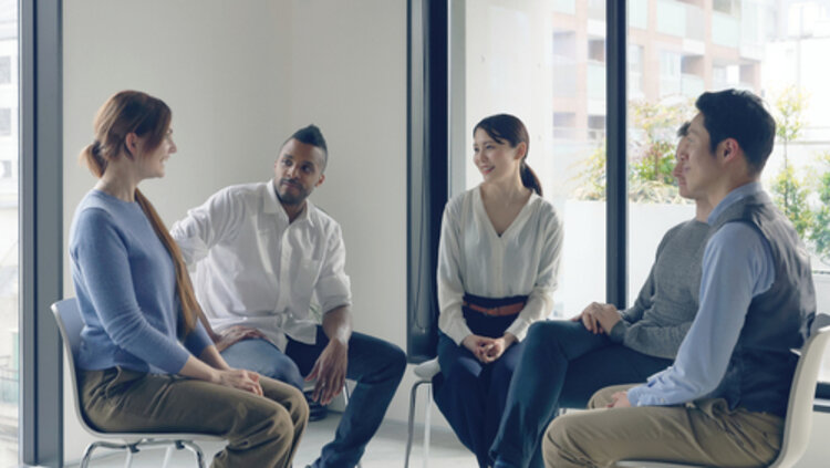 Menschen verschiedener Herkunft sitzen im Halbkreis in einem Raum und unterhalten sich. 