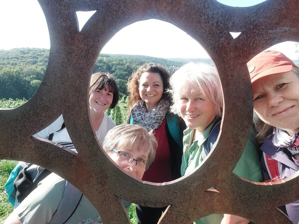Fünf Frauen lächeln für ein gemeinsames Foto durch die runden Aussparungen einer großen Metallskulptur. Im Hintergrund ist eine grüne, hügelige Landschaft zu sehen.