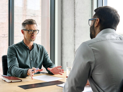 Zwei Männer sitzen sich in einem hellen, modernen Büro mit großen Fenstern gegenüber und führen ein Gespräch. Der Mann links trägt eine dunkle Brille, ein grünes Hemd und hat kurz geschnittene, graumelierte Haare. Er hält einen Kugelschreiber in der Hand und gestikuliert leicht mit der anderen Hand. Der Mann rechts, mit Bart und Brille, trägt ein hellgraues Hemd und hört aufmerksam zu. Auf dem Tisch zwischen ihnen liegen ein Notizbuch, ein Blatt Papier, ein Tablet und ein Glas Wasser. Im Hintergrund sind hohe Fenster mit Blick auf eine Stadtlandschaft zu sehen. Die Atmosphäre wirkt professionell und konzentriert.