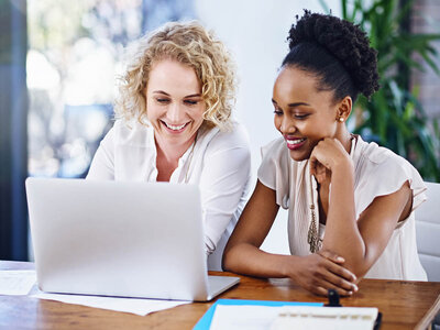 Zwei Frauen sitzen an einem Tisch und arbeiten gemeinsam an einem Laptop. Die Frau links, mit lockigen blonden Haaren und einem weißen Hemd, lächelt und schaut auf den Bildschirm. Die Frau rechts, mit hochgesteckten dunklen Haaren und einem cremefarbenen Hemd, stützt ihr Kinn auf die Hand und scheint ebenfalls auf den Laptop zu schauen. Beide wirken freundlich und engagiert in ihrer Arbeit.