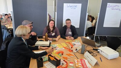 Eine Gruppe von vier Personen sitzt an einem Tisch in einer Seminarumgebung. Sie tragen Namensschilder und lächeln. Im Hintergrund sind Flipcharts mit den Überschriften ‚Wünsche mit IdS‘ und ‚Zukunftsperspektive' zu sehen. Auf dem Tisch liegen Informationsmaterialien, Stifte, ein Laptop und ein Beamer.