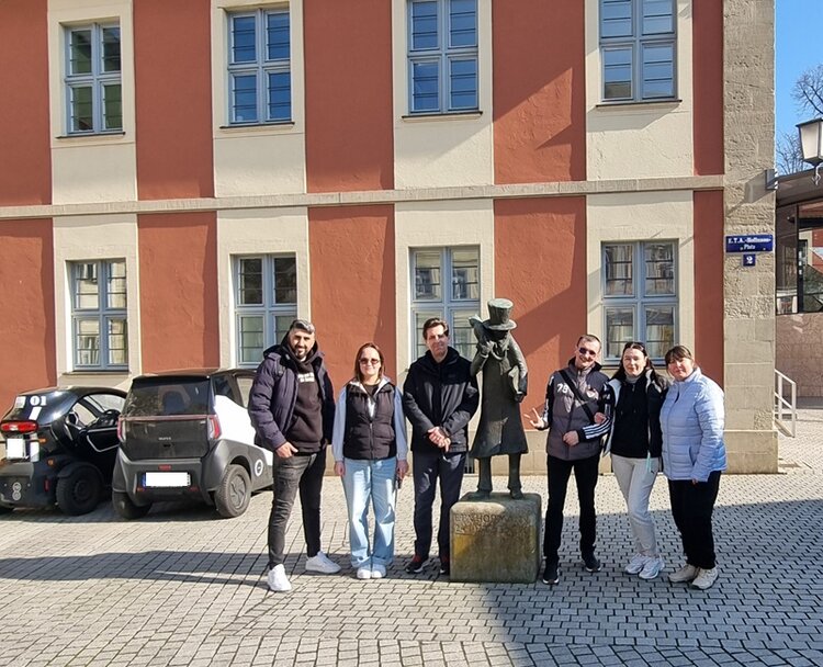 Die Teilnehmenden der Euro-Schulen Bamberg stehen nebeneinander auf einem gepflasterten Platz vor einem historischen Gebäude mit rotbrauner Fassade und großen Fenstern. In der Mitte der Gruppe befindet sich eine bronzene Statue auf einem Sockel. Im Hintergrund sind zwei geparkte Autos zu sehen.