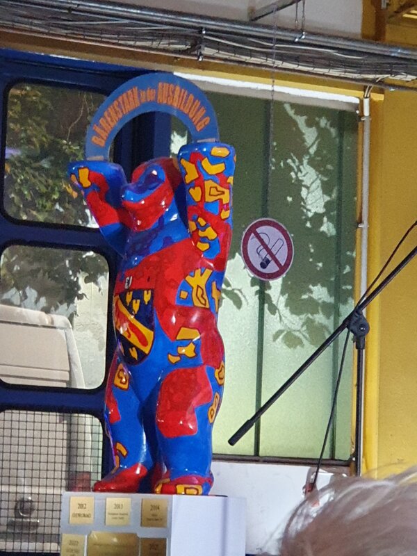 Eine Bärenfigur, die in den Farben Gelb, Blau und Rot angemalt ist und das Stadtwappen von Berlin-Reinickendorf auf der Brust hat, steht vor einer Glasfront.