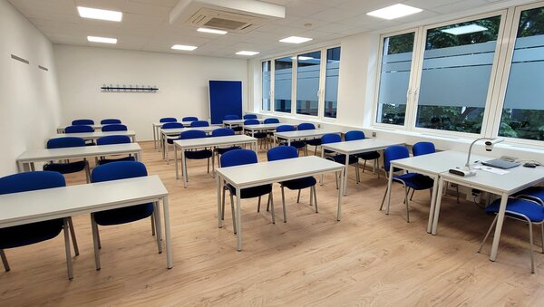 Zweiter Unterrichtsraum mit vielen weißen Tischen und blauen Stühlen, großer Fensterfront rechts und blauem Pinnboard vorne.