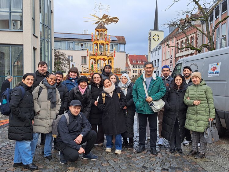 Eine Gruppe von Menschen steht gemeinsam auf einem Marktplatz vor einer großen, beleuchteten Weihnachtspyramide, umgeben von Stadtgebäuden und Kirchturm bei winterlichem Tageslicht.