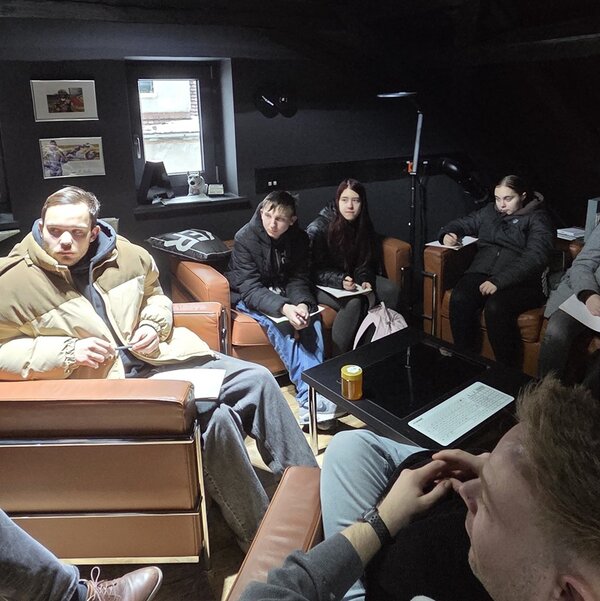 Mehrere Auszubildende der Euro-Schulen Hoyerswerda sitzen in einer modernen Werbeagentur auf braunen Ledersesseln und Sofas um einen schwarzen Couchtisch. Die Gruppe hält Notizblöcke und Stifte in den Händen, einige machen sich Notizen. Auf dem Tisch liegen Papierunterlagen und eine Getränkedose. Die Wände sind dunkel gestrichen, an einer Wand hängen gerahmte Bilder. Durch ein Fenster fällt Tageslicht in den Raum. Die Atmosphäre wirkt konzentriert und ruhig, der Fokus liegt auf dem gemeinsamen Lernen und dem Austausch während des Besuchs in der Werbeagentur.