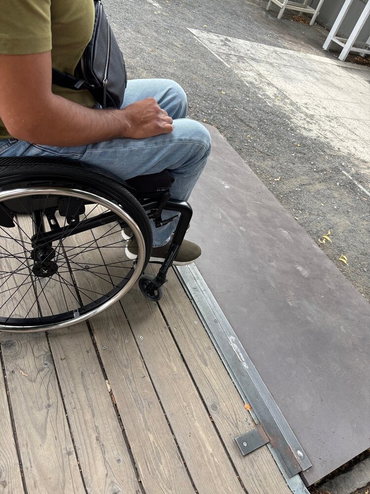 Ein Rollstuhlfahrer mit blauer Jeans und olivgrünem T-Shirt befindet sich auf einer hölzernen Plattform vor einer Metallrampe. Die Rampe verbindet die Holzfläche mit dem angrenzenden Asphaltboden. Im Hintergrund sind ein weißer Pfeil auf dem Boden und einige Metallstrukturen zu sehen.