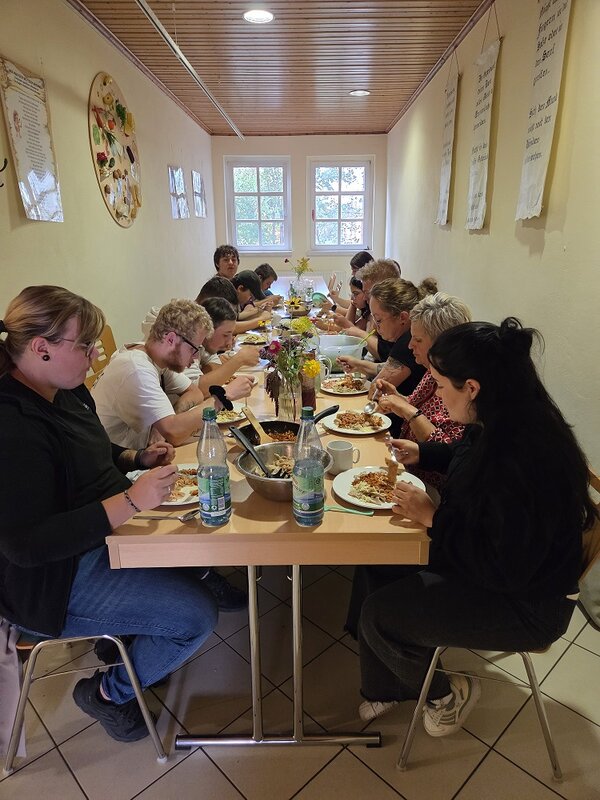 Eine Gruppe von Menschen sitzt an einem langen Holztisch in einem hellen Raum mit Holzvertäfelung und isst gemeinsam. Auf dem Tisch stehen Teller mit Essen, Wasserflaschen und eine Schüssel mit Salat. An den Wänden hängen Plakate.