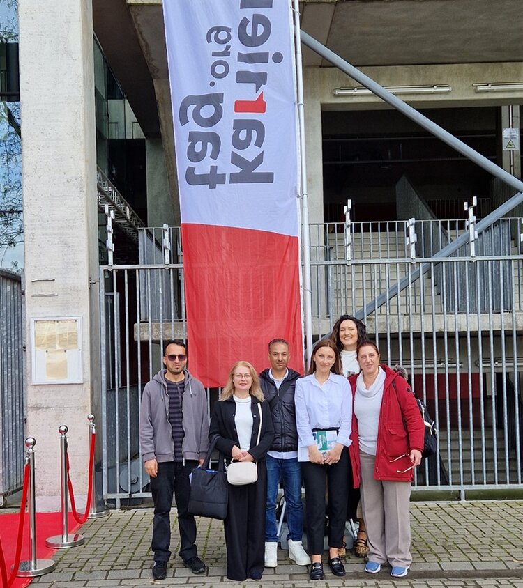 Vor einem Gebäude mit Betonfassade und Metallzaun steht eine Gruppe von Teilnehmenden der Euro-Schulen Kerpen. Im Hintergrund hängt ein großes, vertikal ausgerichtetes Banner mit weiß-rotem Farbfeld und klar lesbarer Schrift. Links sind rote Absperrbänder und silberne Ständer zu sehen. Die Personen tragen unterschiedliche Jacken und halten Taschen oder Gegenstände in den Händen.