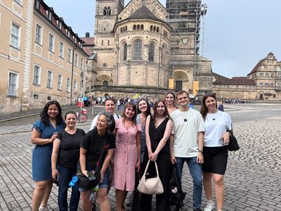 Gruppenfoto der gut gelaunten Teilnehmenden auf einem gepflasterten Platz vor einem historischen Gebäude mit romanischen Rundbogenfenstern und einer großen, teilweise eingerüsteten Fassade in Bamberg. Die Personen sind unterschiedlich gekleidet, einige tragen helle, andere dunkle Kleidung. Im Hintergrund sind weitere Menschen und Gebäude mit hellen Fassaden zu sehen.
