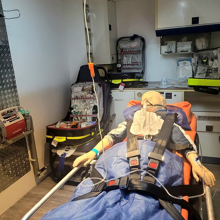 Eine Puppe liegt auf einer Trage festgeschnallt in einem Raum, der einem Krankenwagen ähnelt. Sie trägt eine Sauerstoffmaske und ist an eine Infusion angeschlossen. Um sie herum stehen mehrere Notfallkoffer mit medizinscher Ausrüstung.