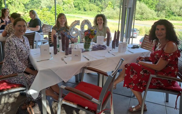 Vier lächelnde Frauen sitzen an einem festlich gedeckten Tisch in einem hellen Raum mit Blick auf einen Garten. Auf dem Tisch steht in der Mitte eine silberne '60' als Dekoration sowie Blumen, Gläser und Geschenktüten.