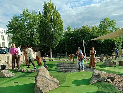 Mehrere Personen spielen auf einem Minigolfplatz mit künstlichem Rasen und verschiedenen Hindernissen wie großen Steinen und Baumstämmen. Die Spieler sind konzentriert und halten Putter in den Händen. Der Platz ist von einem Zaun umgeben, und im Hintergrund sind Bäume und ein Gebäude zu erkennen.