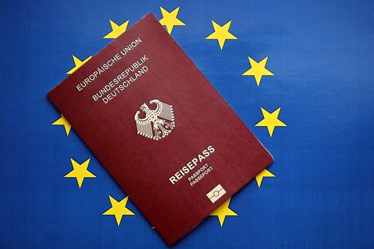 Ein bordeauxroter Reisepass mit dem Bundesadler und goldener Aufschrift liegt schräg auf einer Europaflagge mit gelbem Sternenkreis auf blauem Grund. Die Aufschrift auf dem Pass lautet: 'EUROPÄISCHE UNION BUNDESREPUBLIK DEUTSCHLAND REISEPASS'. Unten ist ein goldfarbenes Passsymbol zu sehen.