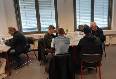 Mehrere Teilnehmende sitzen in einem Seminarraum an Tischen zusammen und führen Gespräche bei einer Informations- oder Beratungsveranstaltung.