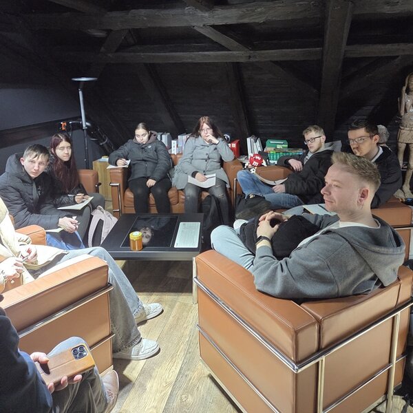 Das Bild zeigt eine Gruppe von Auszubildenden der Euro-Schulen Hoyerswerda, die sich in einer Werbeagentur zu einer Gesprächs- und Arbeitsrunde versammelt haben. Die angehenden Verkäuferinnen und Verkäufer sitzen in bequemen Ledersesseln im Dachgeschoss eines Agenturbüros mit sichtbaren Holzbalken. In der Mitte steht ein niedriger Tisch mit Notizen und Unterlagen. Einige Teilnehmende halten Schreibblöcke oder Stifte in der Hand und machen sich Notizen, andere hören aufmerksam zu. Die ruhige, konzentrierte Atmosphäre vermittelt einen praxisnahen Einblick in Marketing, Kommunikation und kreative Arbeitsprozesse im Rahmen der beruflichen Ausbildung.