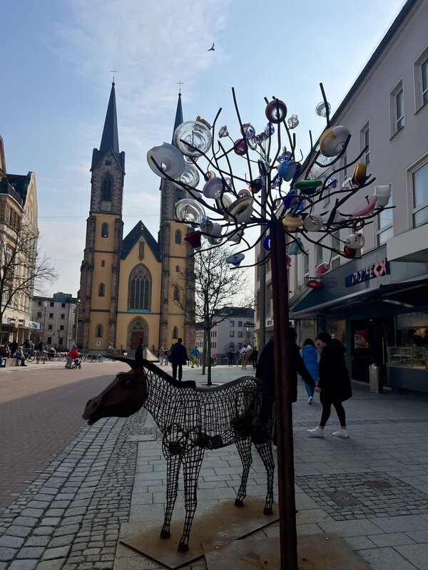Die Innenstadt von Hof. Zu sehen ist eine Gasse, an deren Ende die gelbe Marienkirche steht. Im Vordergrund stehen zwei Kunstwerke: Ein Esel aus Metall und daneben ein Baum aus Metall, an dessen Äste alte Lampenschirme aus Glas hängen.