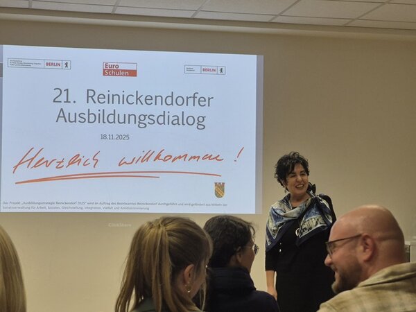 Eine Frau steht vor einer groß projizierten Begrüßungsfolie mit der Aufschrift ‚21. Reinickendorfer Ausbildungsdialog – Herzlich willkommen!‘ und spricht zu den anwesenden Gästen. Im Vordergrund sind mehrere Zuhörer von hinten zu sehen. Die Atmosphäre wirkt freundlich und dialogorientiert. Das Foto entstand im Rahmen des 21. Reinickendorfer Ausbildungsdialogs an den Euro-Schulen Berlin.