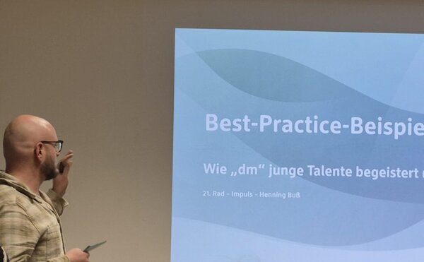 Ein Mann steht vor einer Präsentationsfolie und hält einen Vortrag mit dem Titel ‚Best-Practice-Beispiele – Wie dm junge Talente begeistert‘. Der Sprecher zeigt mit der Hand auf die Folie, während mehrere Teilnehmer im Publikum zuhören. Die Szene spielt in einem modernen Seminarraum beim 21. Reinickendorfer Ausbildungsdialog an den Euro-Schulen Berlin.