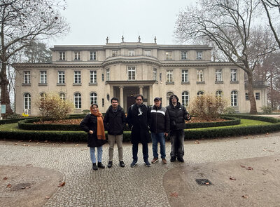 Fünf Personen posieren auf einem Kiesweg vor einer hellen, repräsentativen Villa mit vielen Fenstern; der kreisförmig angelegte Garten mit geschnittenen Hecken und kahlen Bäumen lässt auf einen kühlen Wintertag schließen.