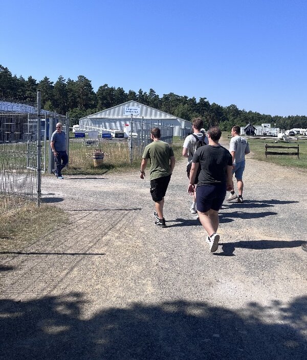 Eine kleine Gruppe geht über das Gelände eines Flugplatzes in Richtung eines Hangars und weiterer Gebäude.