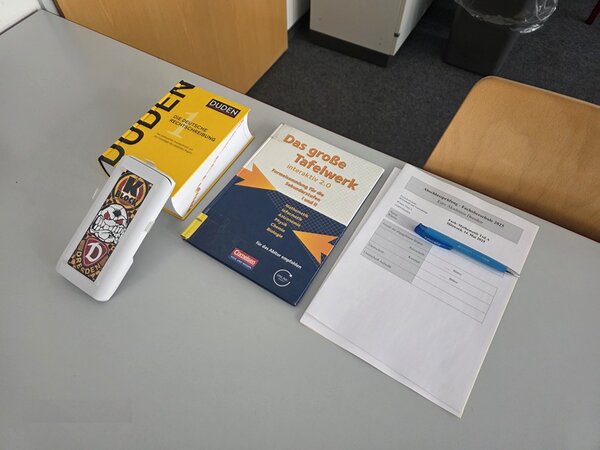 Auf einem weißen Tisch liegt ein Deutsch-Duden, ein Kugelschreiber, Arbeitsblätter zut Prüfung sowie ein Lehrbuch.