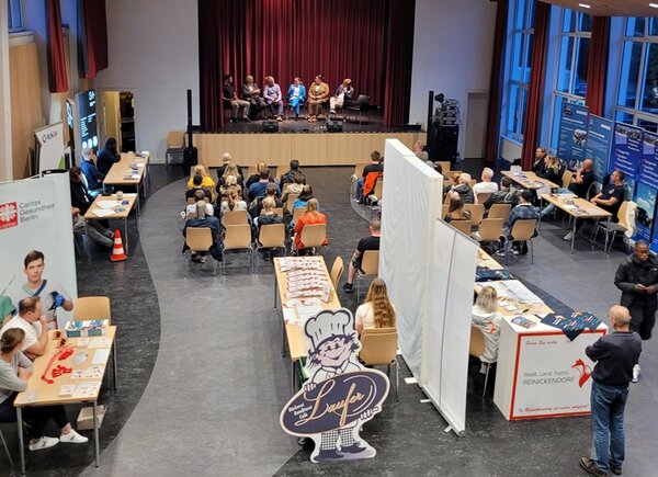 Teilnehmende sitzen auf der Bühne und diskutieren, das Publikum hört zu, Informationsstände stehen bereit. 